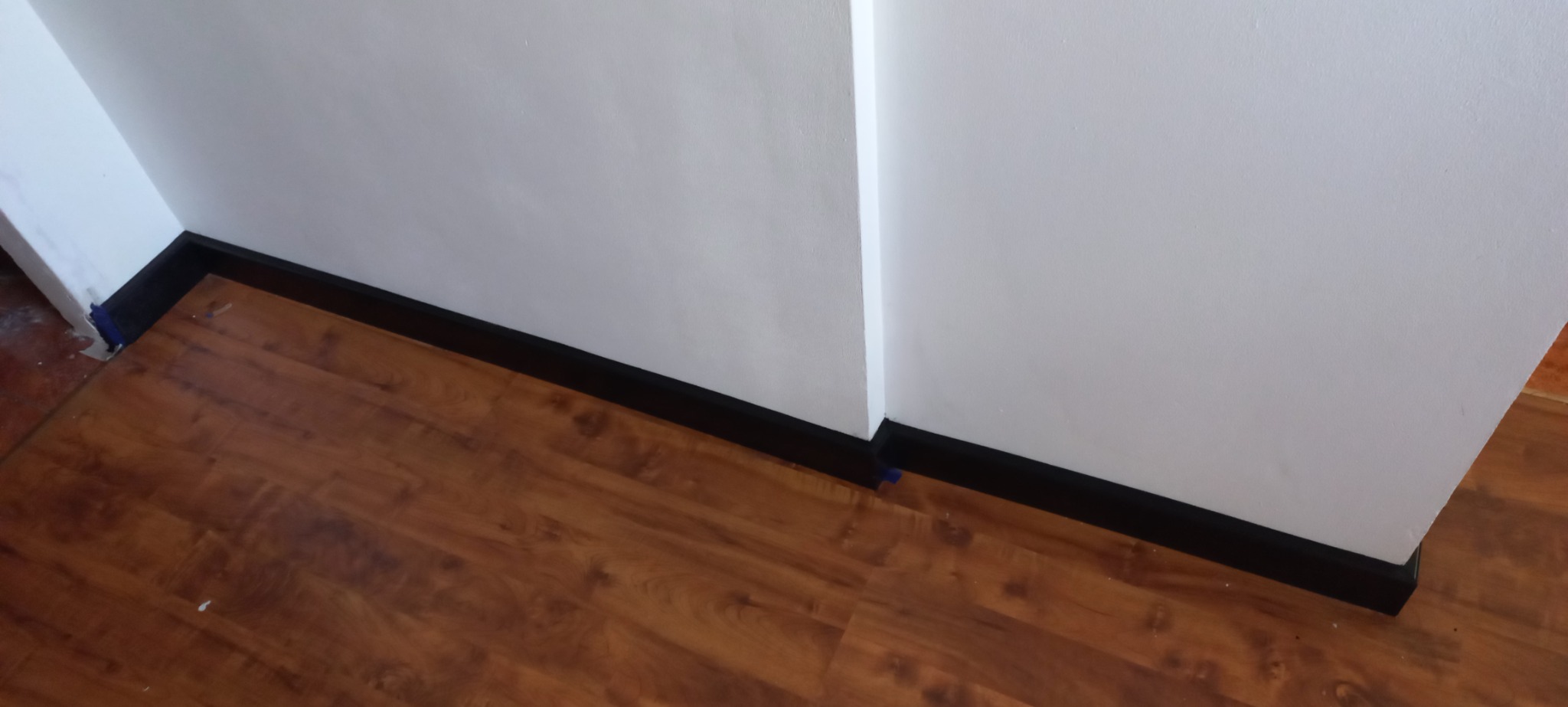 Brązowe panele podłogowe z czarną listwą przypodłogową przy białej ścianie, zabezpieczone niebieską taśmą malarską w rogu.