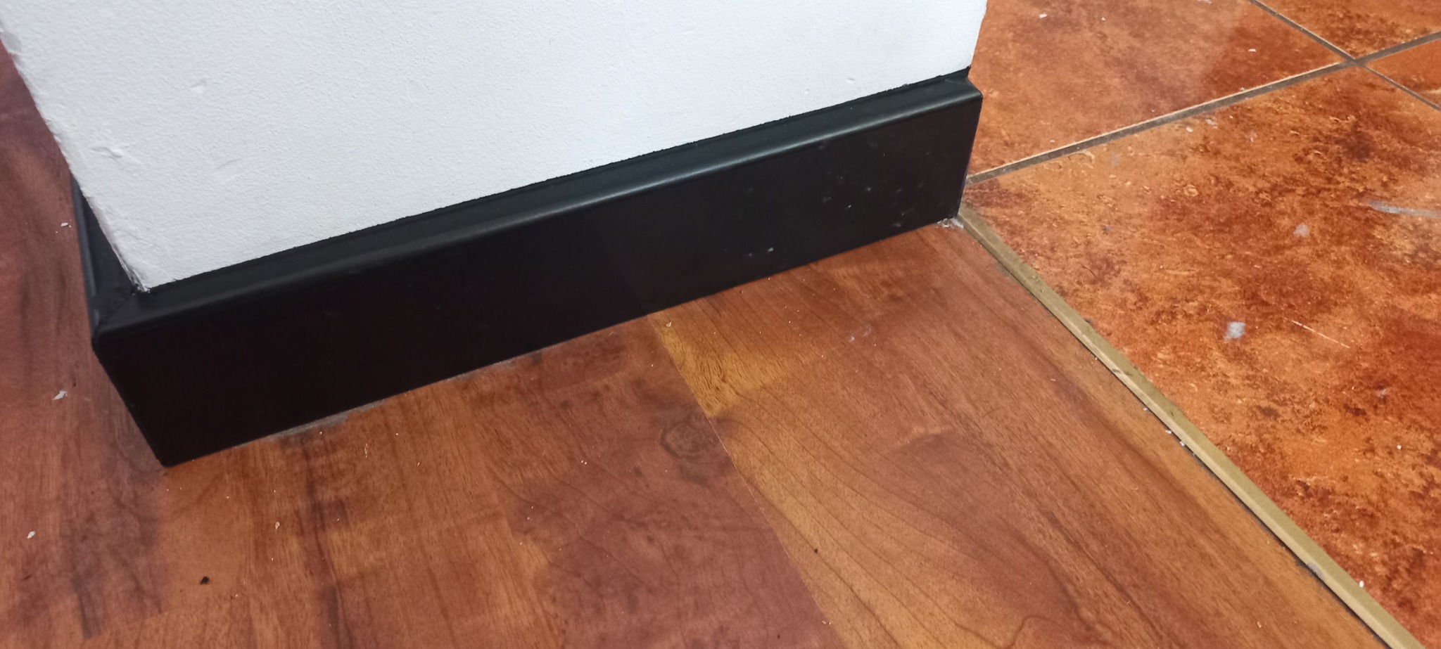 Połączenie podłóg: panele imitujące drewno i terakota oddzielone złotą listwą, czarna listwa przypodłogowa przy białej ścianie.