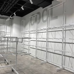 KAPKIP ALIAKSEI KAMORNIK - Wnętrze sklepu z szarymi płytkami podłogowymi i białymi, metalowymi regałami na ubrania zamontowanymi na ścianie, z napisem COOL w tle.