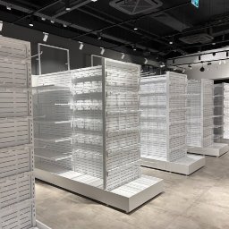 KAPKIP ALIAKSEI KAMORNIK - Białe, metalowe regały sklepowe z perforowanymi ściankami i haczykami, ustawione w rzędach na szarych płytkach podłogowych, pod czarnym sufitem z widocznymi instalacjami i oświetleniem punktowym.