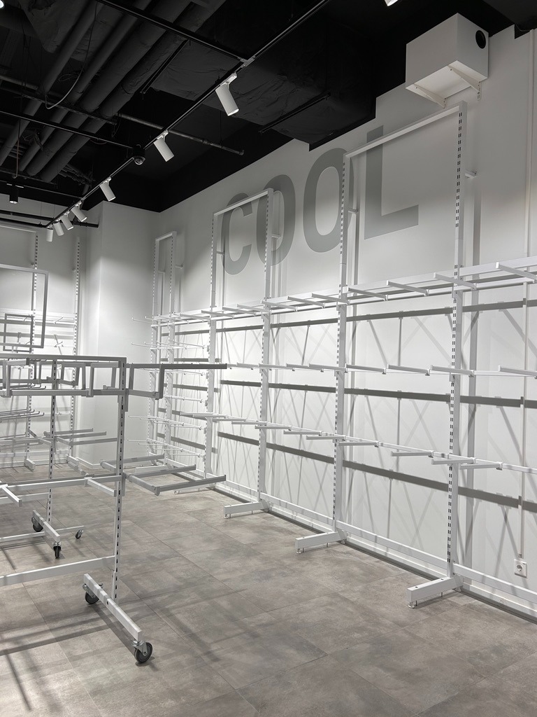 Wnętrze sklepu z szarymi płytkami podłogowymi i białymi, metalowymi regałami na ubrania zamontowanymi na ścianie, z napisem COOL w tle.