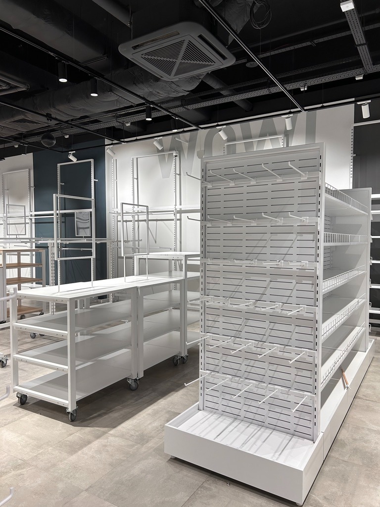 Białe, metalowe regały sklepowe z haczykami i półkami, ustawione na szarej podłodze w jasnym wnętrzu sklepu przed otwarciem.