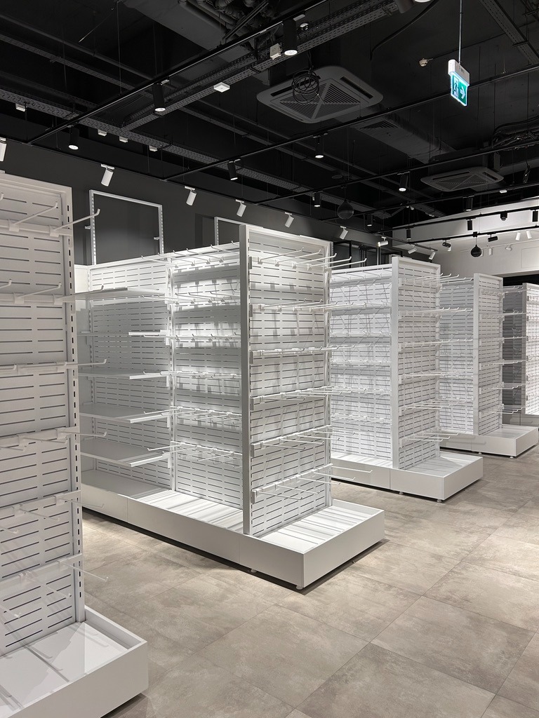 Białe, metalowe regały sklepowe z perforowanymi ściankami i haczykami, ustawione w rzędach na szarych płytkach podłogowych, pod czarnym sufitem z widocznymi instalacjami i oświetleniem punktowym.