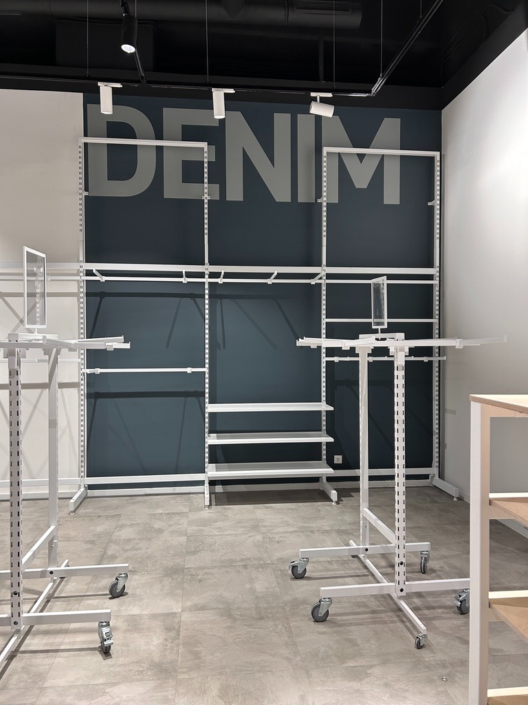 Montaż białych, metalowych regałów sklepowych na kółkach, z półkami i wieszakami, na tle ściany w kolorze denim z napisem DENIM, oświetlenie punktowe na suficie.