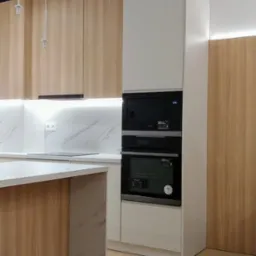 Nowoczesna kuchnia z drewnianymi frontami szafek, białymi elementami i zabudowanym piekarnikiem oraz mikrofalówką, oświetlona listwami LED.