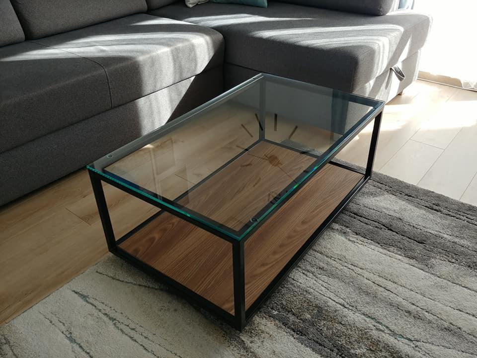 Stolik kawowy z czarną metalową ramą, szklanym blatem i półką z drewna w odcieniach brązu, ustawiony na wzorzystym dywanie obok szarej sofy.