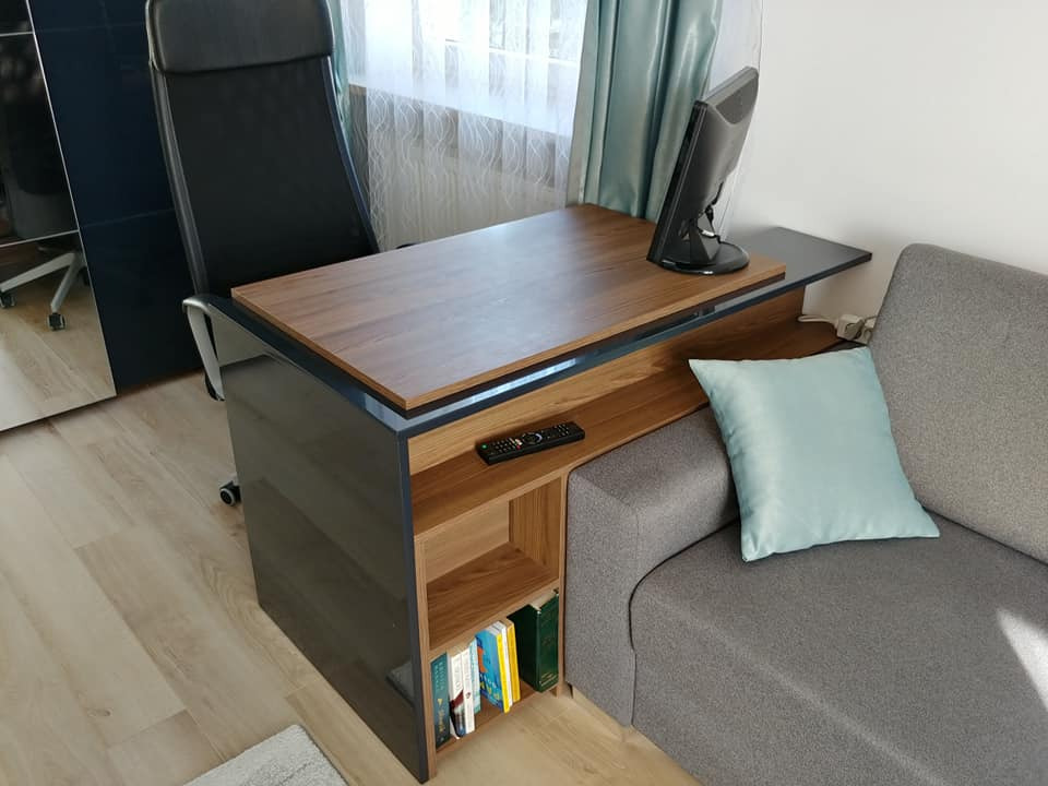 Nowoczesne biurko zintegrowane z regałem i sofą w pokoju dziennym, z blatem w odcieniu drewna, czarnymi błyszczącymi bokami i otwartymi półkami na książki, obok szara sofa z błękitną poduszką.