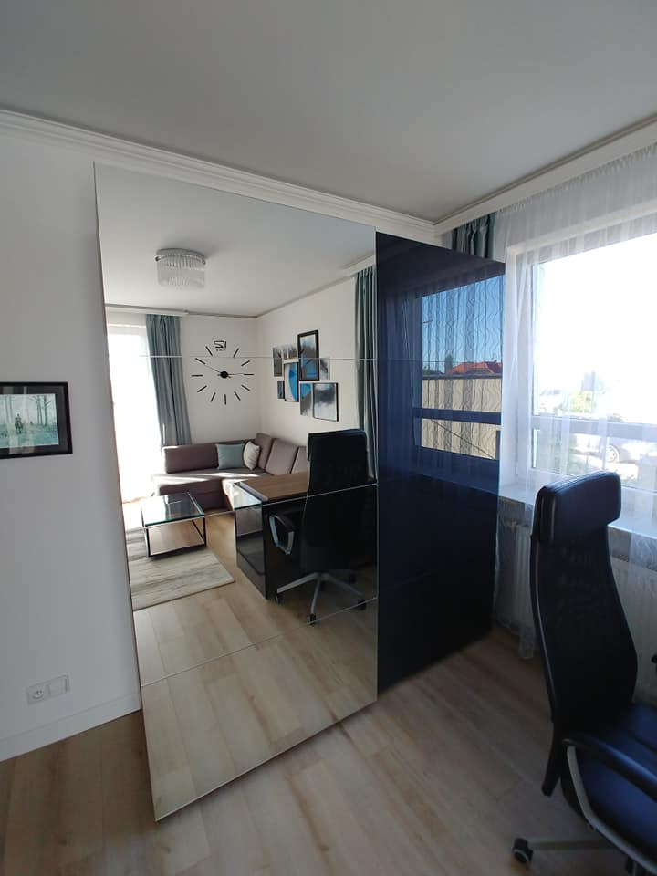 Nowoczesna garderoba z lustrzanymi drzwiami przesuwnymi i czarnym panelem bocznym w jasnym pokoju z widocznym biurkiem i krzesłem.