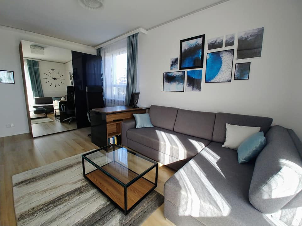 Szafa wnękowa z lustrzanymi drzwiami w salonie w Kańkowie, odbijająca biurko i krzesło, nad szarą sofą abstrakcyjne obrazy.