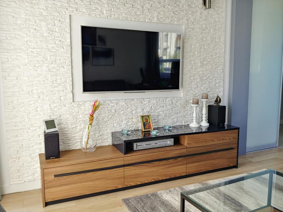 Drewniana szafka RTV z czarnymi uchwytami pod wiszącym telewizorem na ścianie z białej cegły dekoracyjnej, uzupełniona dekoracjami i sprzętem audio.