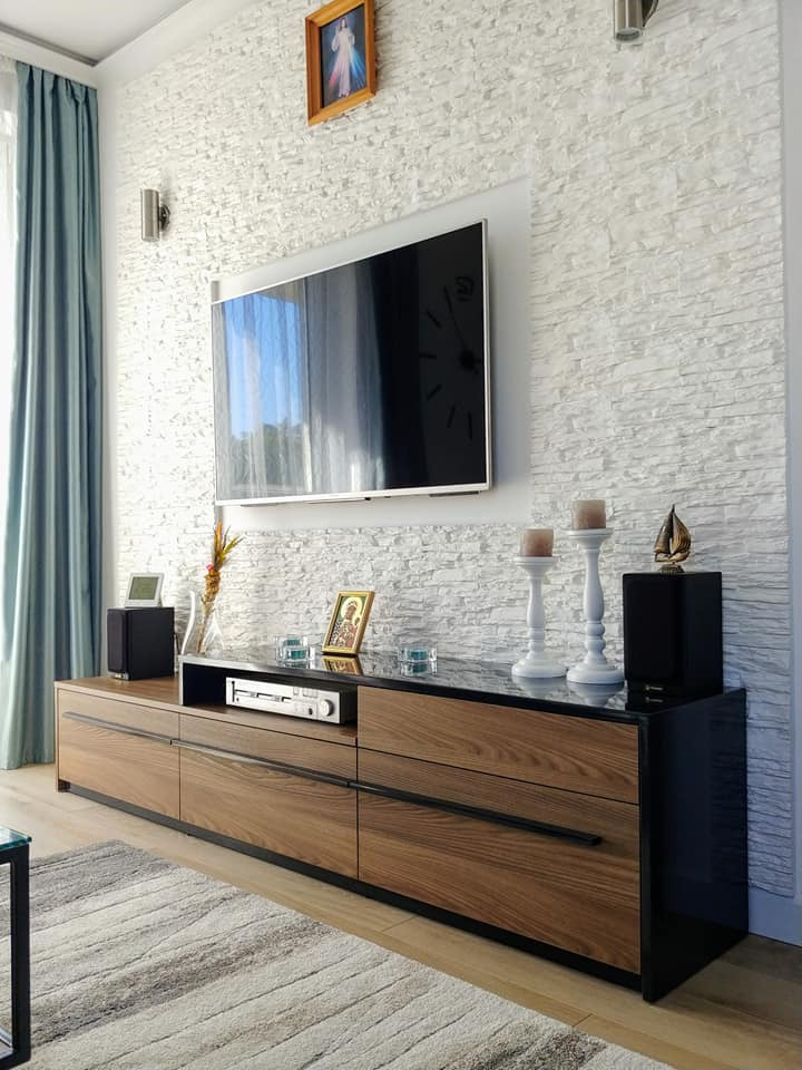 Nowoczesna szafka RTV z drewnianymi frontami szuflad i czarnym blatem, uzupełniona o sprzęt audio, świeczniki i dekoracje, na tle ściany z białej cegły.
