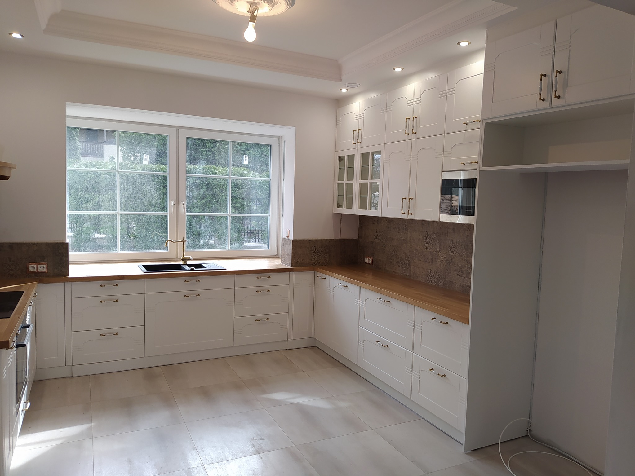 Jasna kuchnia z białymi szafkami w stylu klasycznym, drewniany blat, złota bateria przy oknie, płytki podłogowe w odcieniach szarości.