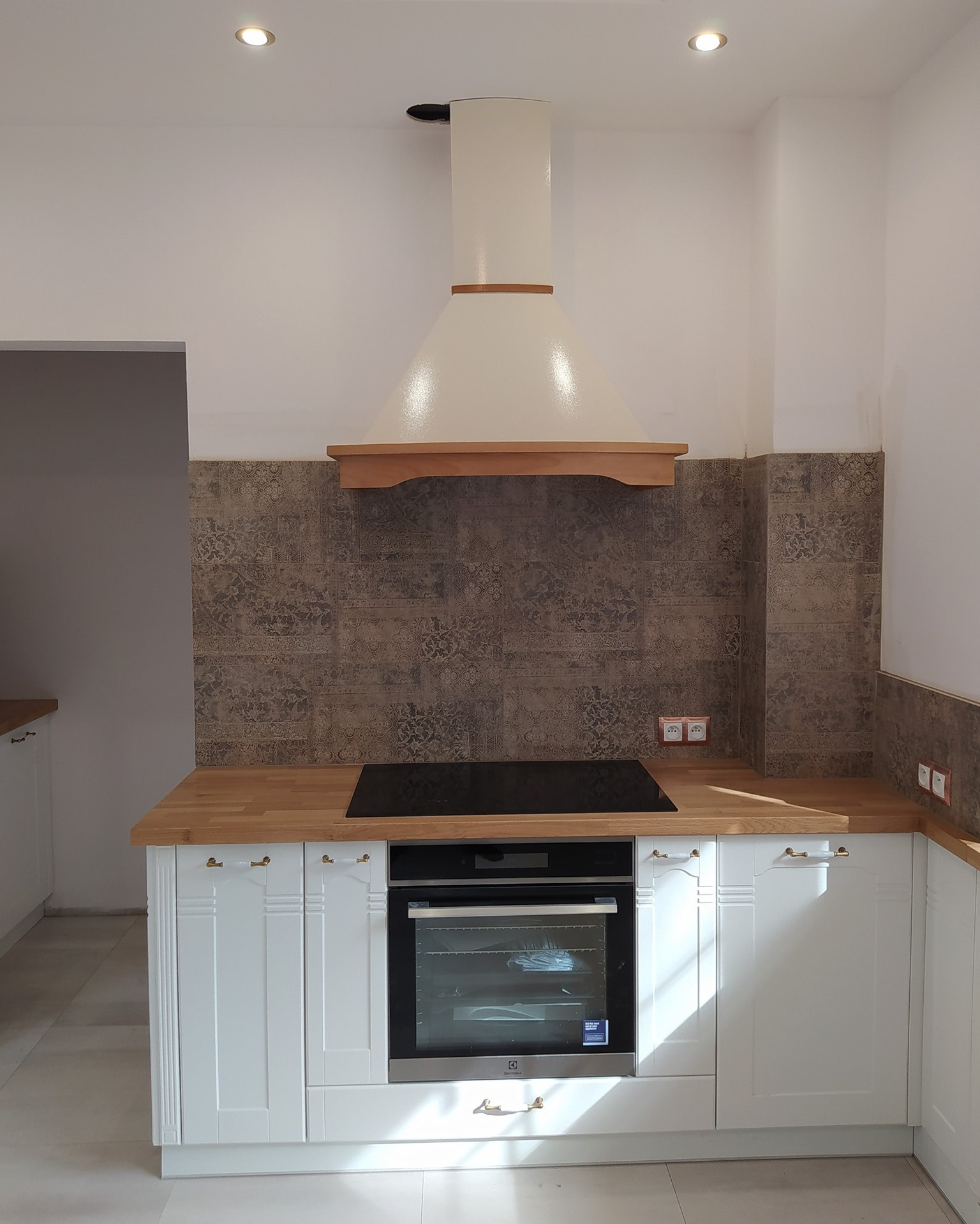 Fragment nowej kuchni z białymi szafkami z frezowanymi frontami i złotymi uchwytami, drewnianym blatem, brązowymi płytkami nad blatem i kremowym okapem z drewnianym wykończeniem.