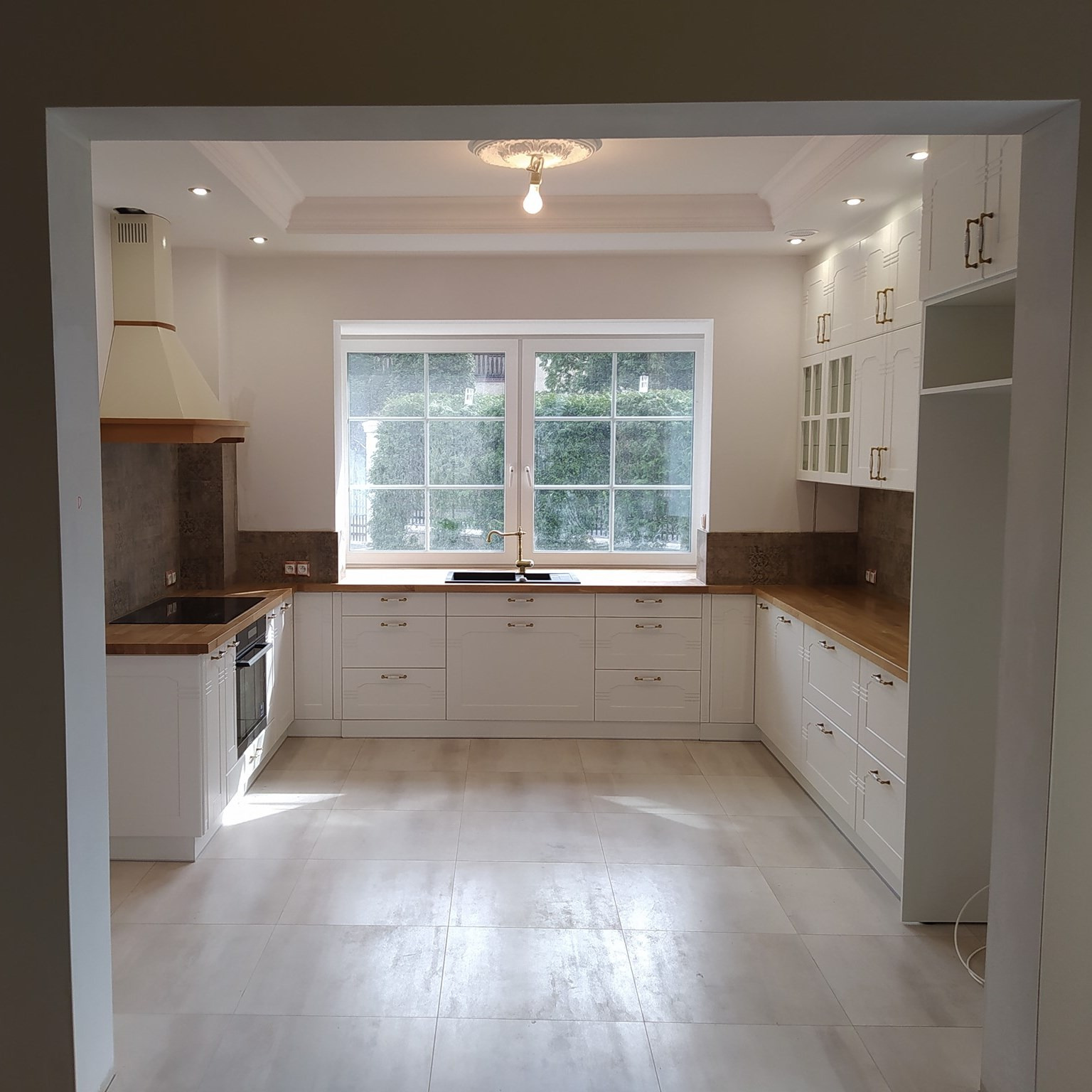 Jasna, przestronna kuchnia z białymi szafkami, drewnianym blatem i dużym oknem, widok z perspektywy wejścia do pomieszczenia.