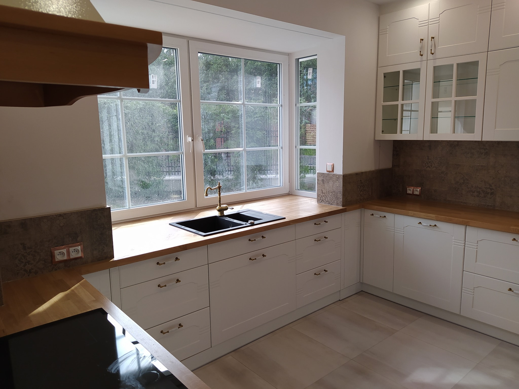 Jasna kuchnia na wymiar z drewnianym blatem, czarnym zlewem pod oknem i białymi frontami szafek z ozdobnymi uchwytami.