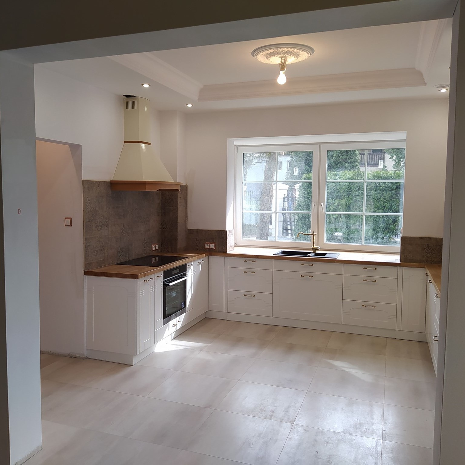 Jasna kuchnia z białymi szafkami, drewnianym blatem i beżowym okapem, widok na ogród przez okno.