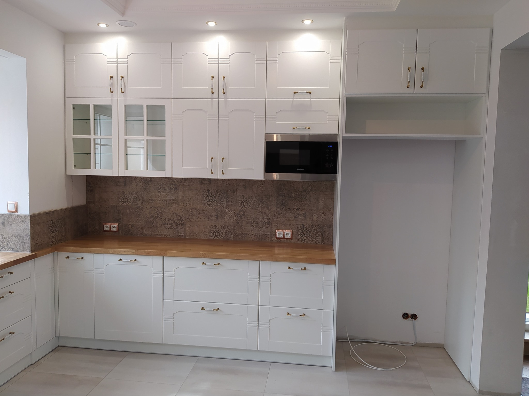 Jasna kuchnia z białymi szafkami z dekoracyjnymi uchwytami, drewnianym blatem i zabudowaną mikrofalówką, wykończona płytkami imitującymi wzór tapety.