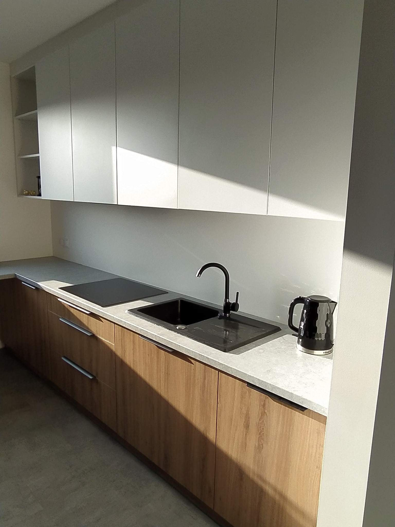 Nowoczesna kuchnia z drewnianymi szafkami dolnymi i białymi szafkami górnymi, czarnym zlewem i czajnikiem, oświetlona popołudniowym słońcem.