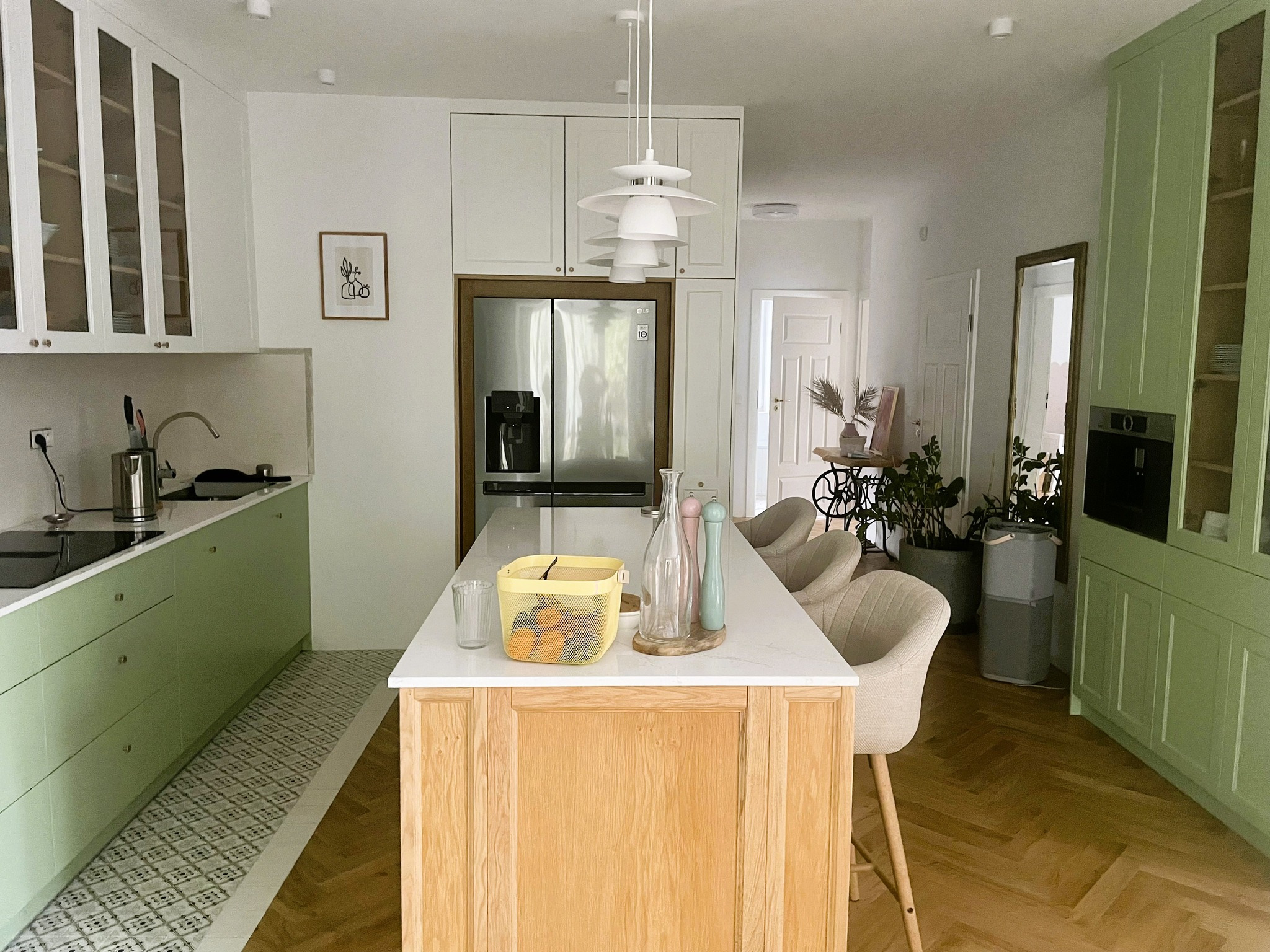 Jasna kuchnia z pistacjowymi szafkami, wyspą z drewnianym frontem i białym blatem, wisząca lampa w stylu skandynawskim.