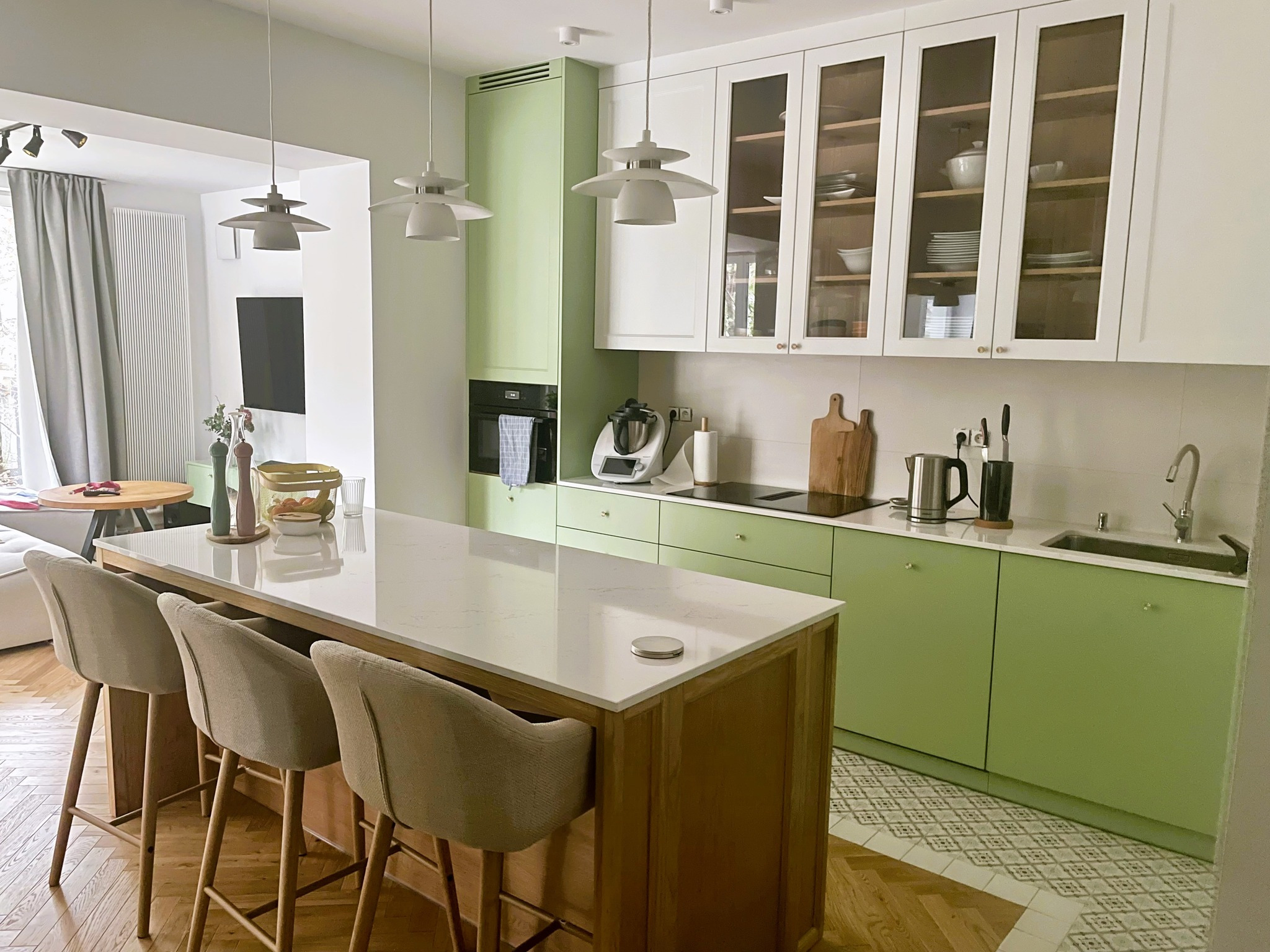 Nowoczesna kuchnia z wyspą, zielonymi szafkami i białymi szafkami wiszącymi z przeszklonymi frontami, oświetlona trzema lampami wiszącymi, z widocznymi barowymi krzesłami i parkietem.