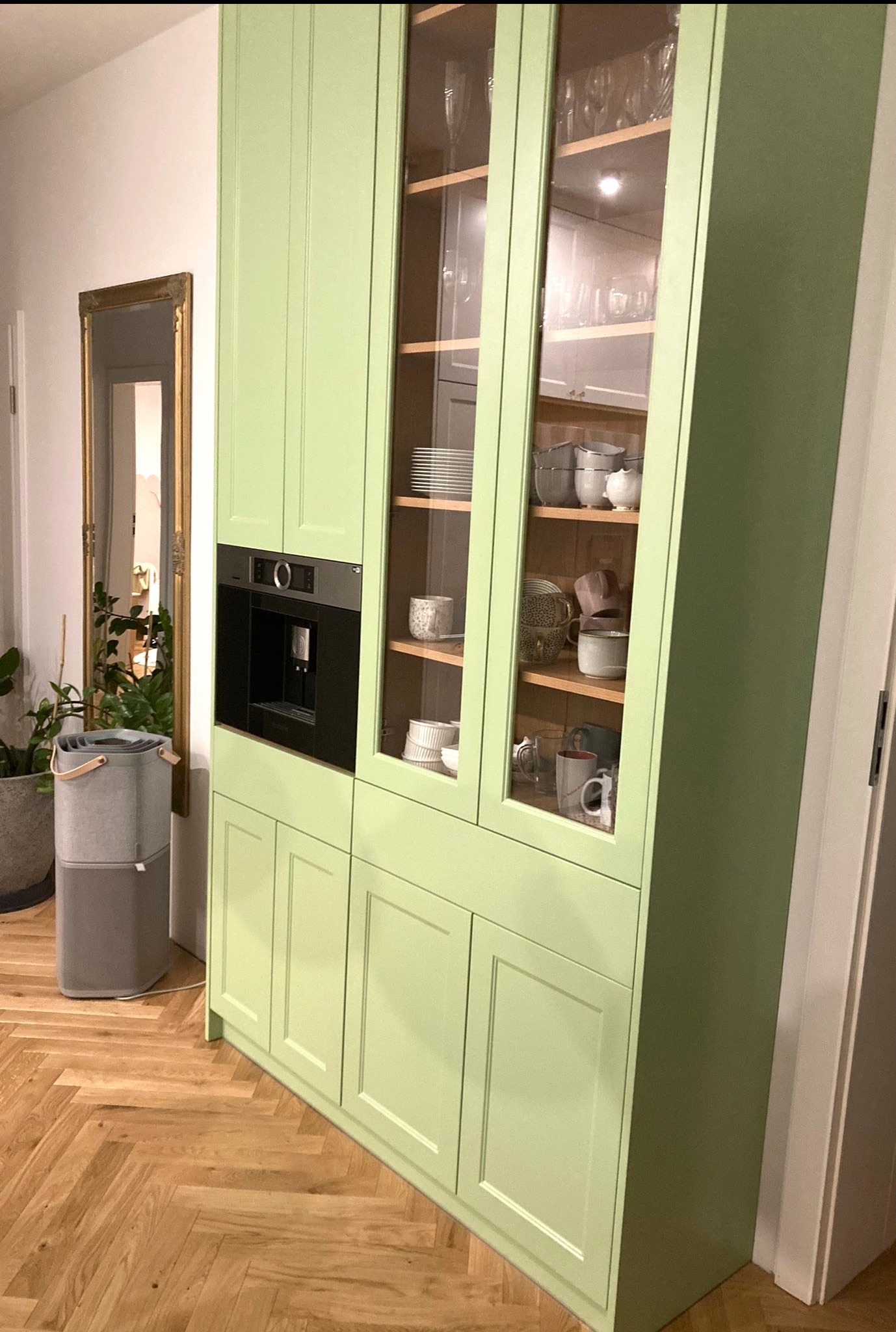 Pistacjowa zabudowa kuchenna z przeszklonymi witrynami i wbudowanym ekspresem do kawy, uzupełniona o parkiet w jodełkę i lustro w złotej ramie.