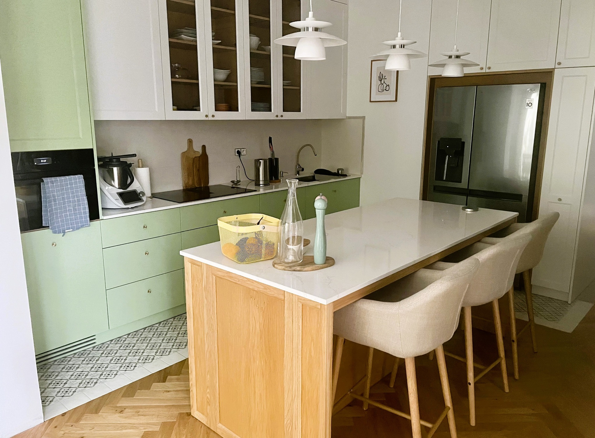 Jasna kuchnia z pistacjowymi szafkami dolnymi, białymi górnymi, wyspą z blatem z konglomeratu i drewnianą podstawą oraz trzema krzesłami barowymi, oświetlona trzema białymi lampami wiszącymi.