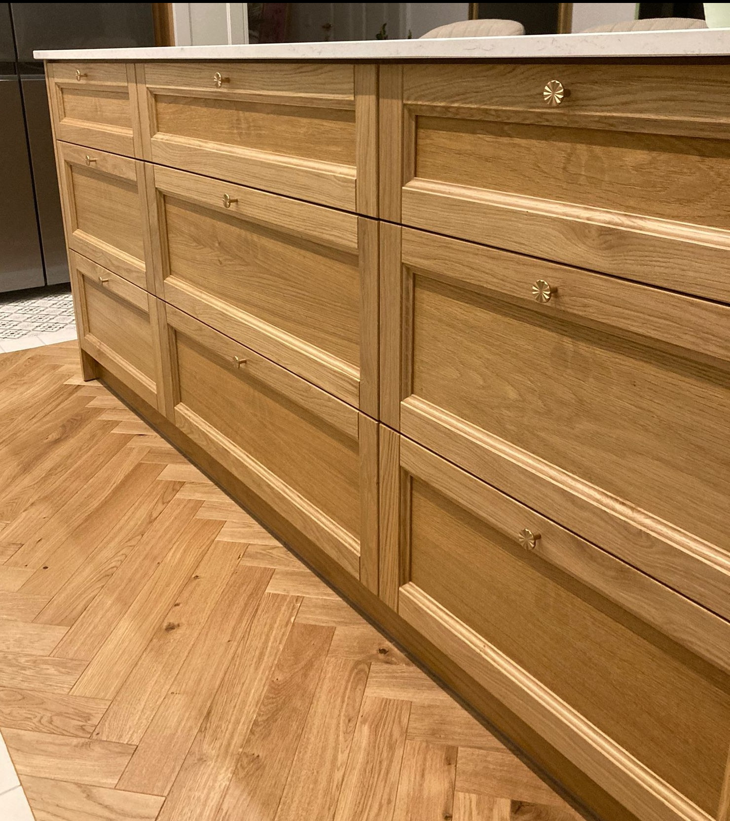 Szczegółowe ujęcie frontu szuflad z jasnego drewna z dekoracyjnymi, złotymi uchwytami na tle parkietu ułożonego w jodełkę.
