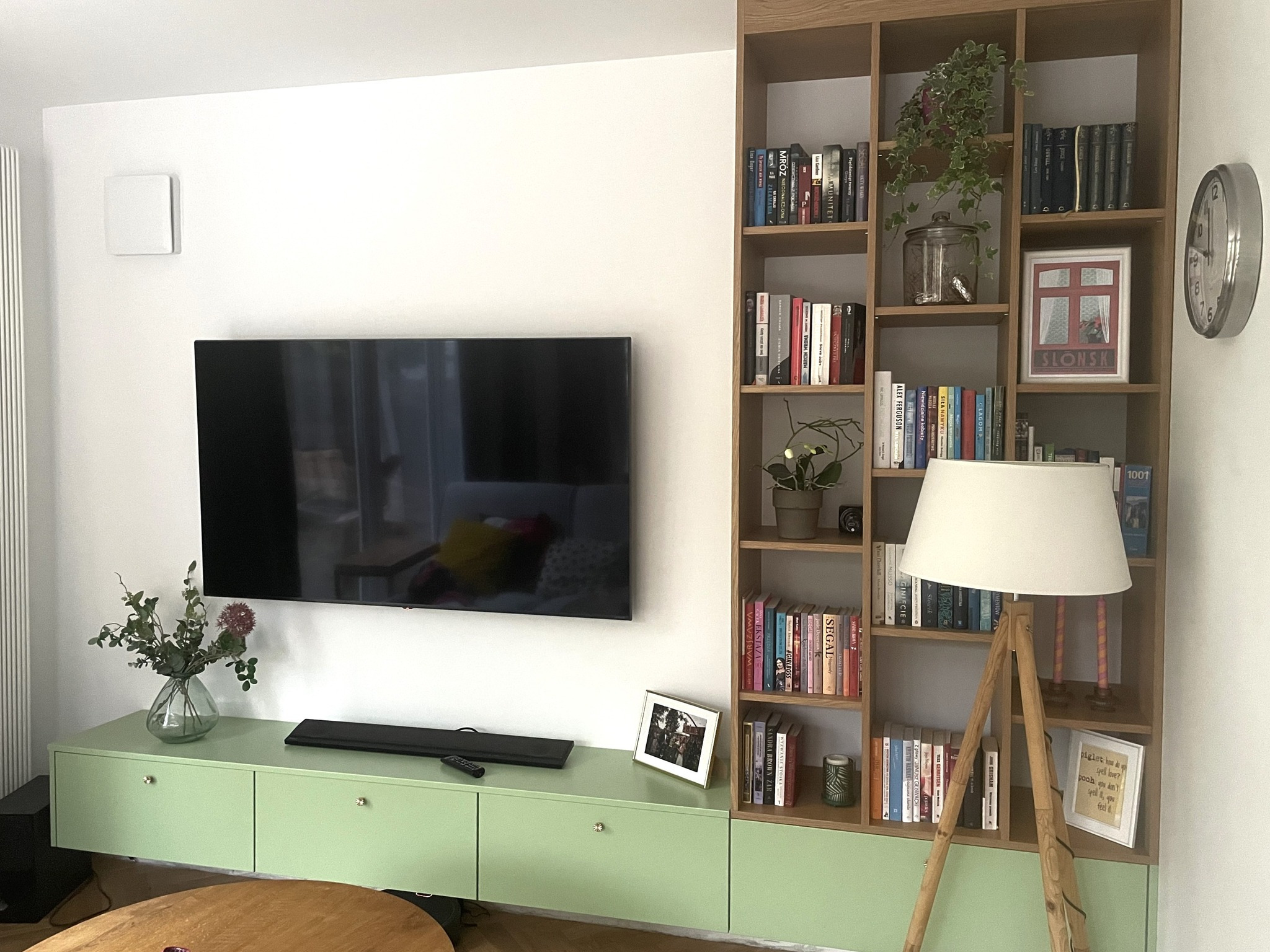 Salon z telewizorem, zieloną szafką RTV i regałem na książki z drewna w ciepłym odcieniu, uzupełniony lampą na trójnogu i dekoracjami.