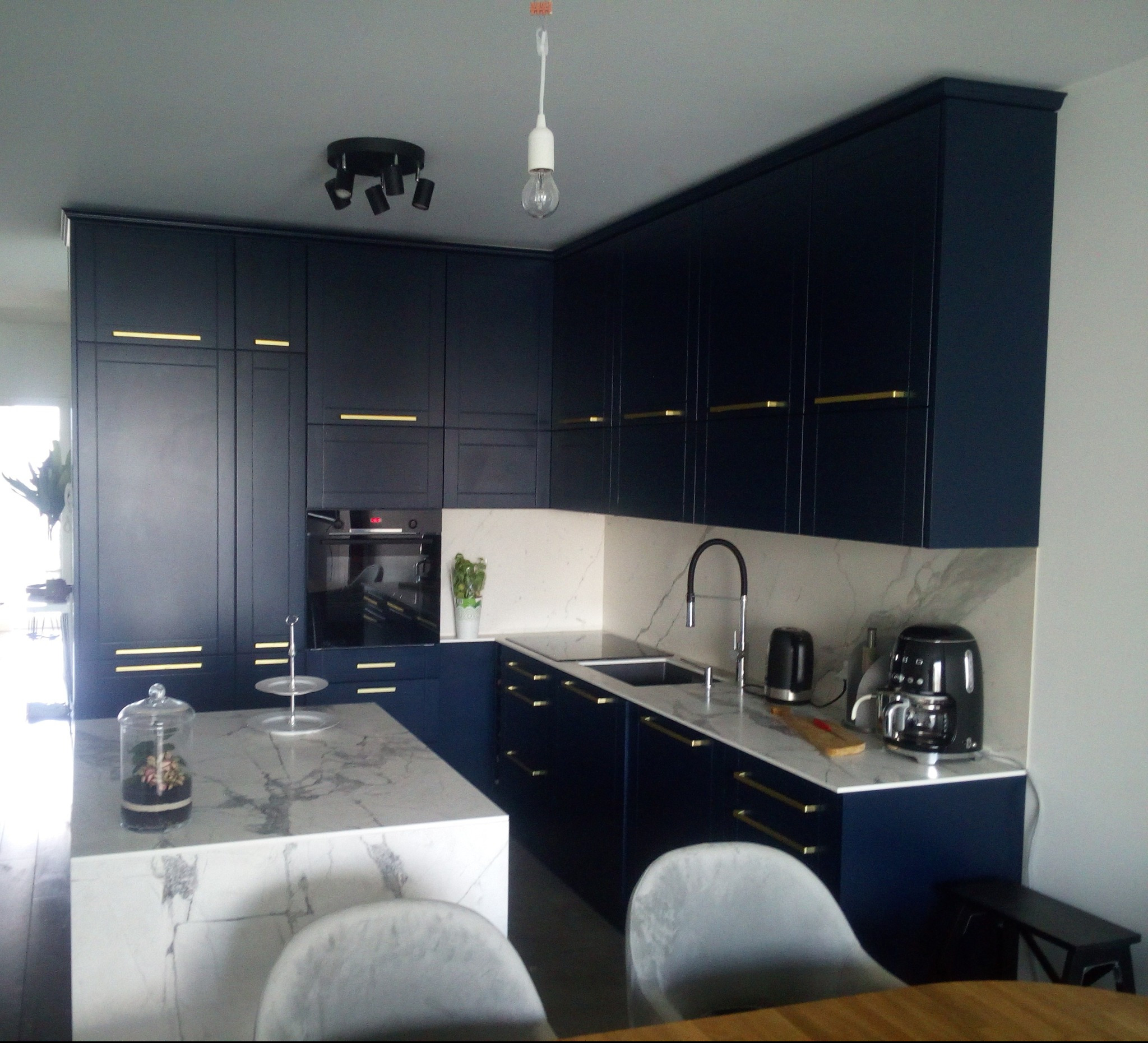 Granatowa zabudowa kuchenna z marmurowym blatem i złotymi uchwytami, wyspa z dekoracjami, oświetlenie sufitowe punktowe i pojedyncza żarówka.