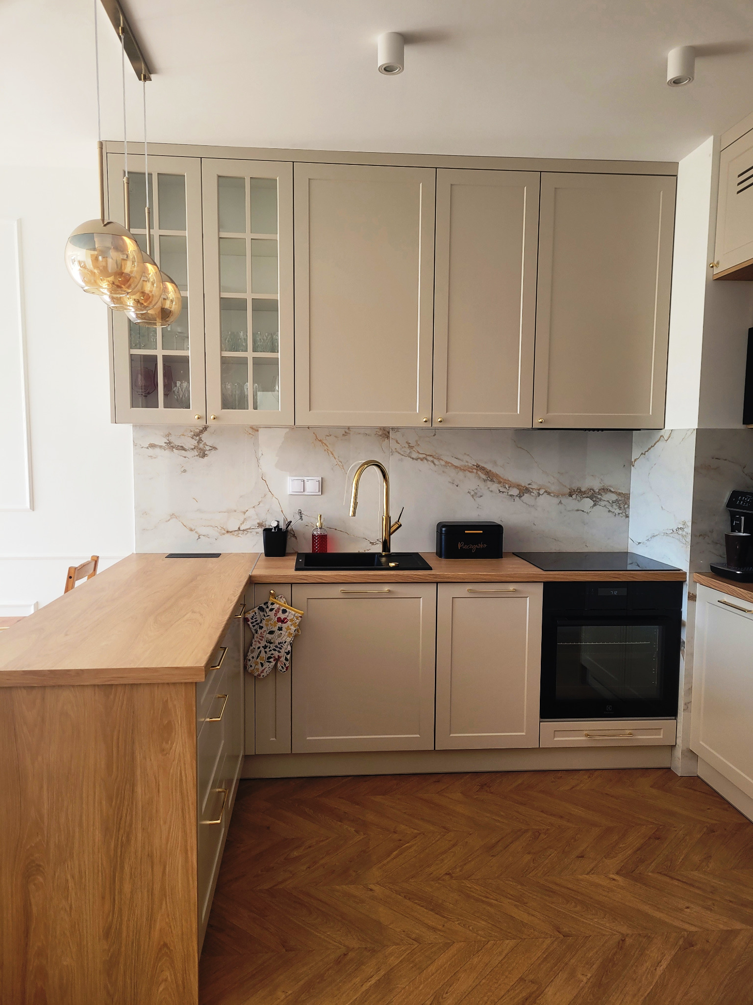 Beżowa zabudowa kuchenna z marmurowym backsplash'em, złotą armaturą i drewnianym blatem, uzupełniona o wiszące, szklane lampy w złotym odcieniu.