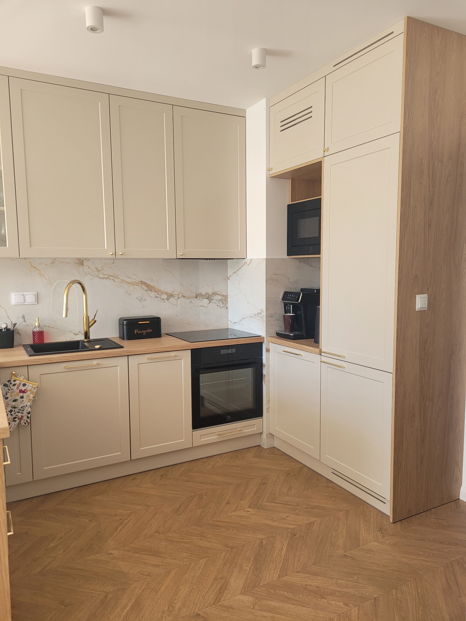 Jasna kuchnia z drewnianą podłogą w jodełkę, złotą armaturą i czarnym zlewem, z zabudowaną kuchenką i piekarnikiem oraz ekspresem do kawy.