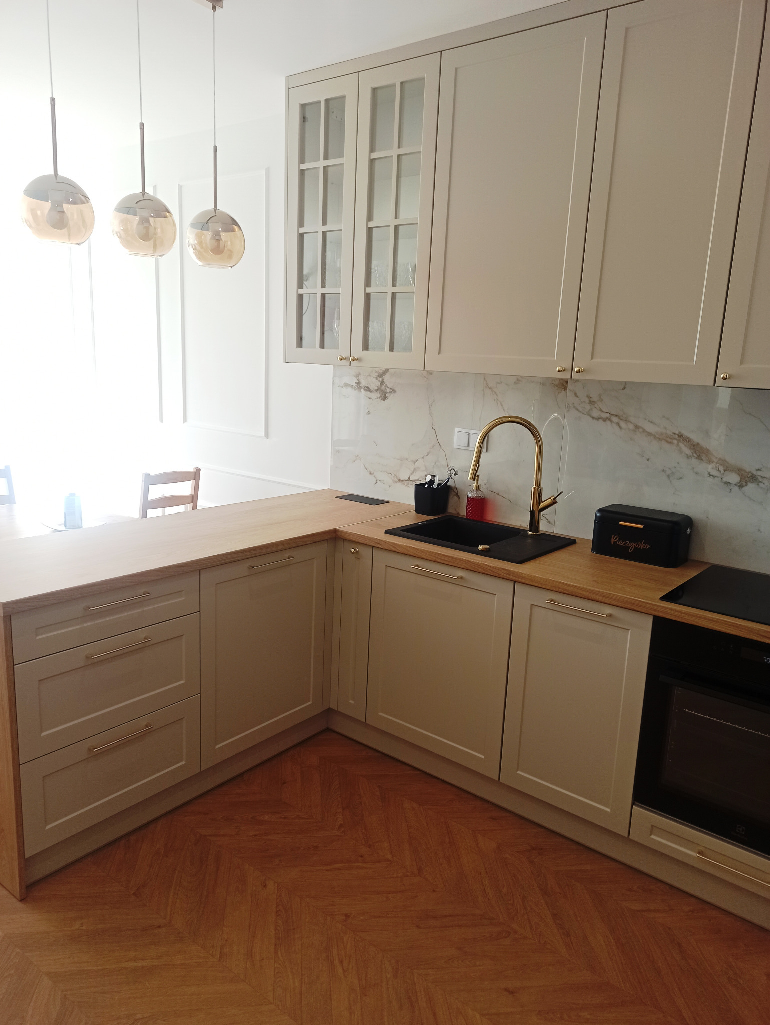 Jasna kuchnia z drewnianym blatem, złotą armaturą i parkietem ułożonym w jodełkę; widoczny czarny zlew i piekarnik.