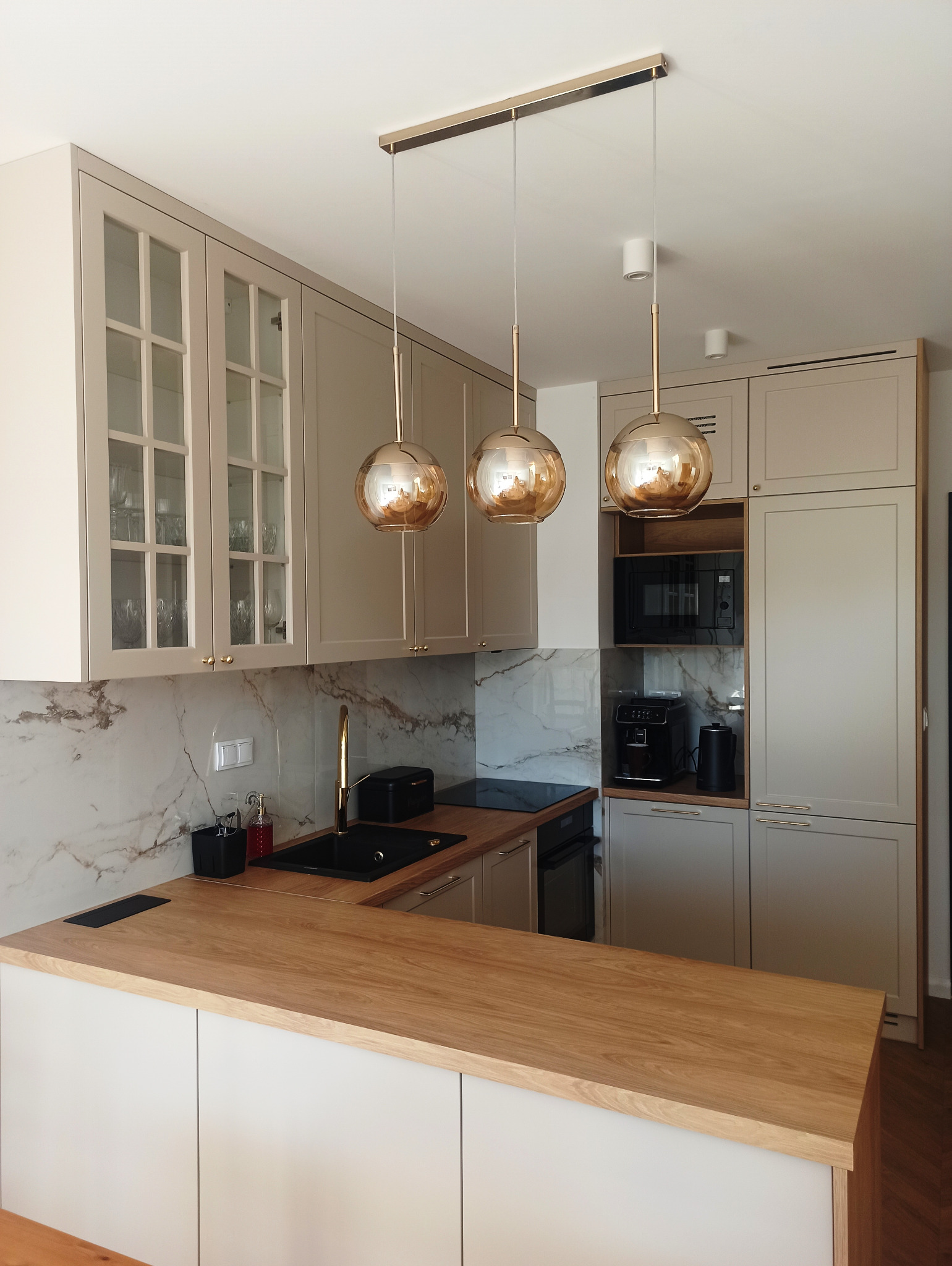 Jasna kuchnia z drewnianym blatem, złotą armaturą i trzema wiszącymi, kulistymi lampami w kolorze miedzi, sprzęt AGD w zabudowie.