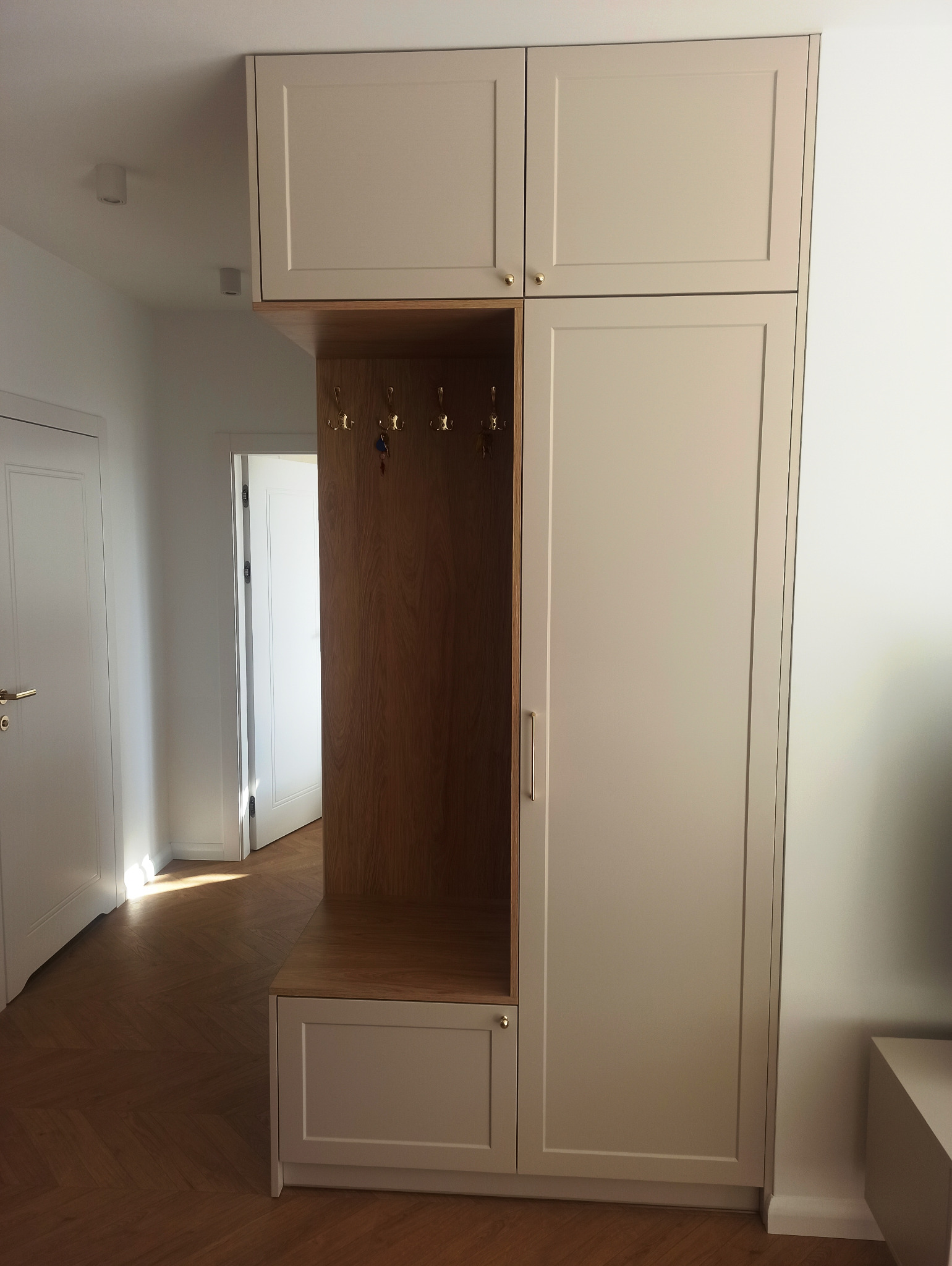 Zabudowa przedpokoju z szafkami w kolorze beżowym, drewnianym panelem z wieszakami i dolną szafką, na podłodze parkiet w jodełkę.