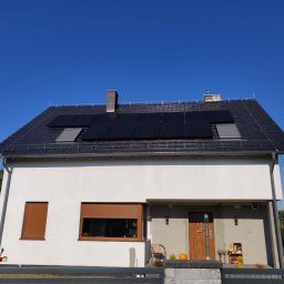 SUNFIT Pakosław Szyszko - Biały dom jednorodzinny z ciemną dachówką i panelami słonecznymi na dachu, widok z przodu, błękitne niebo w tle.