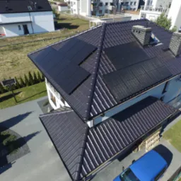 Widok z góry na dom jednorodzinny z brązowym dachem pokrytym panelami słonecznymi; na podjeździe stoi niebieski samochód dostawczy.