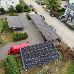 SUNFIT Pakosław Szyszko - Widok z góry na instalację fotowoltaiczną pełniącą funkcję zadaszenia parkingowego, z czerwonym vanem w pobliżu, na tle zabudowy mieszkalnej.