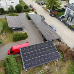 Widok z góry na instalację fotowoltaiczną pełniącą funkcję zadaszenia parkingowego, z czerwonym vanem w pobliżu, na tle zabudowy mieszkalnej.