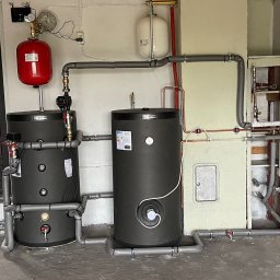 SUNFIT Pakosław Szyszko - Dwa czarne akumulatory ciepła z czerwoną i białą naczyniem wzbiorczym, połączone rurami w kotłowni z widocznymi elementami instalacji.