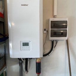 SUNFIT Pakosław Szyszko - Biały, wiszący na ścianie akumulator ciepła marki Auratsu z podłączonymi rurami i filtrem, obok skrzynka elektryczna z przewodami w peszlu.