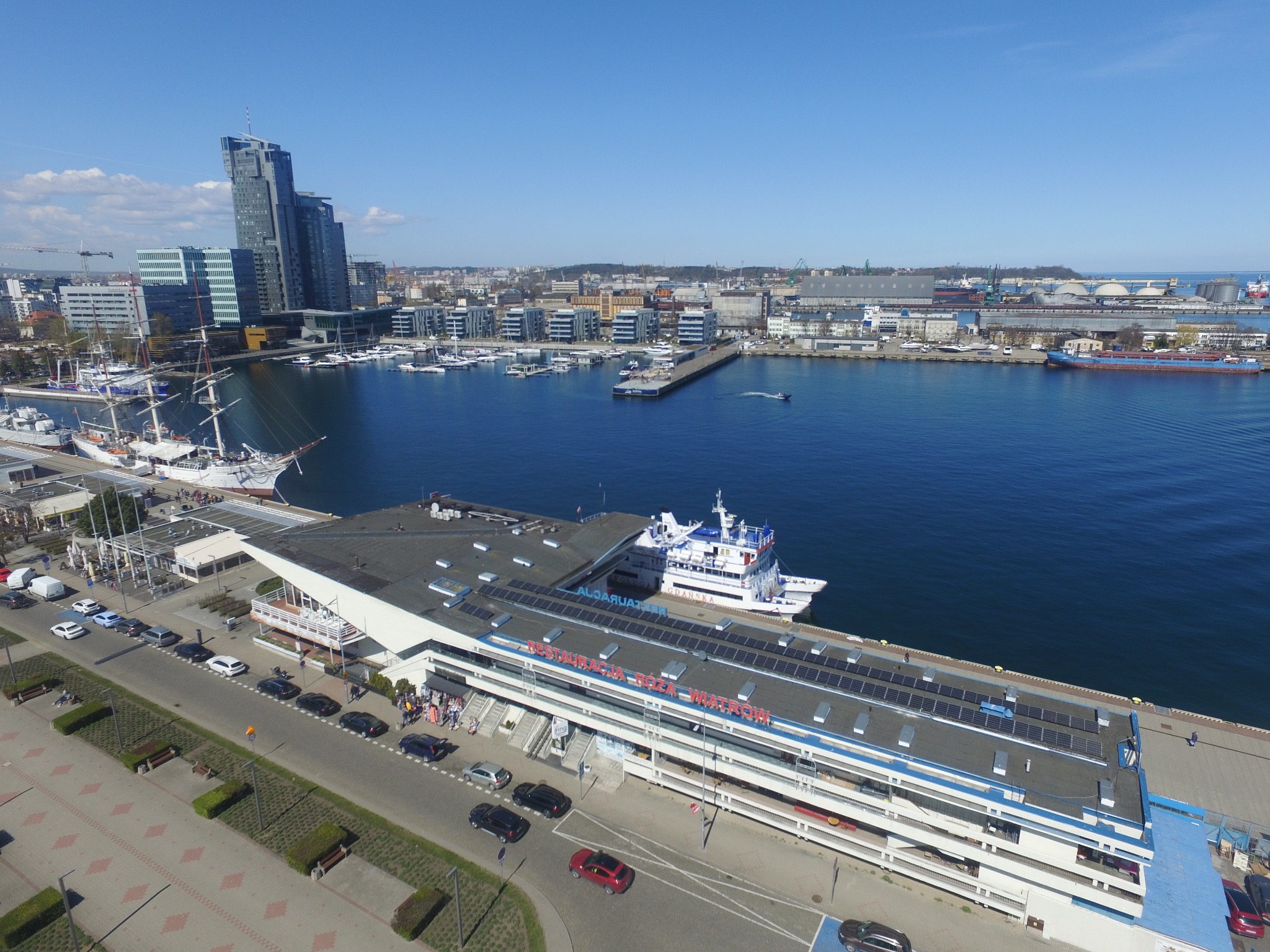 Panoramiczny widok z góry na port w Gdyni z budynkiem z panelami słonecznymi na dachu, jachtami i nowoczesną architekturą w tle, w słoneczny dzień.