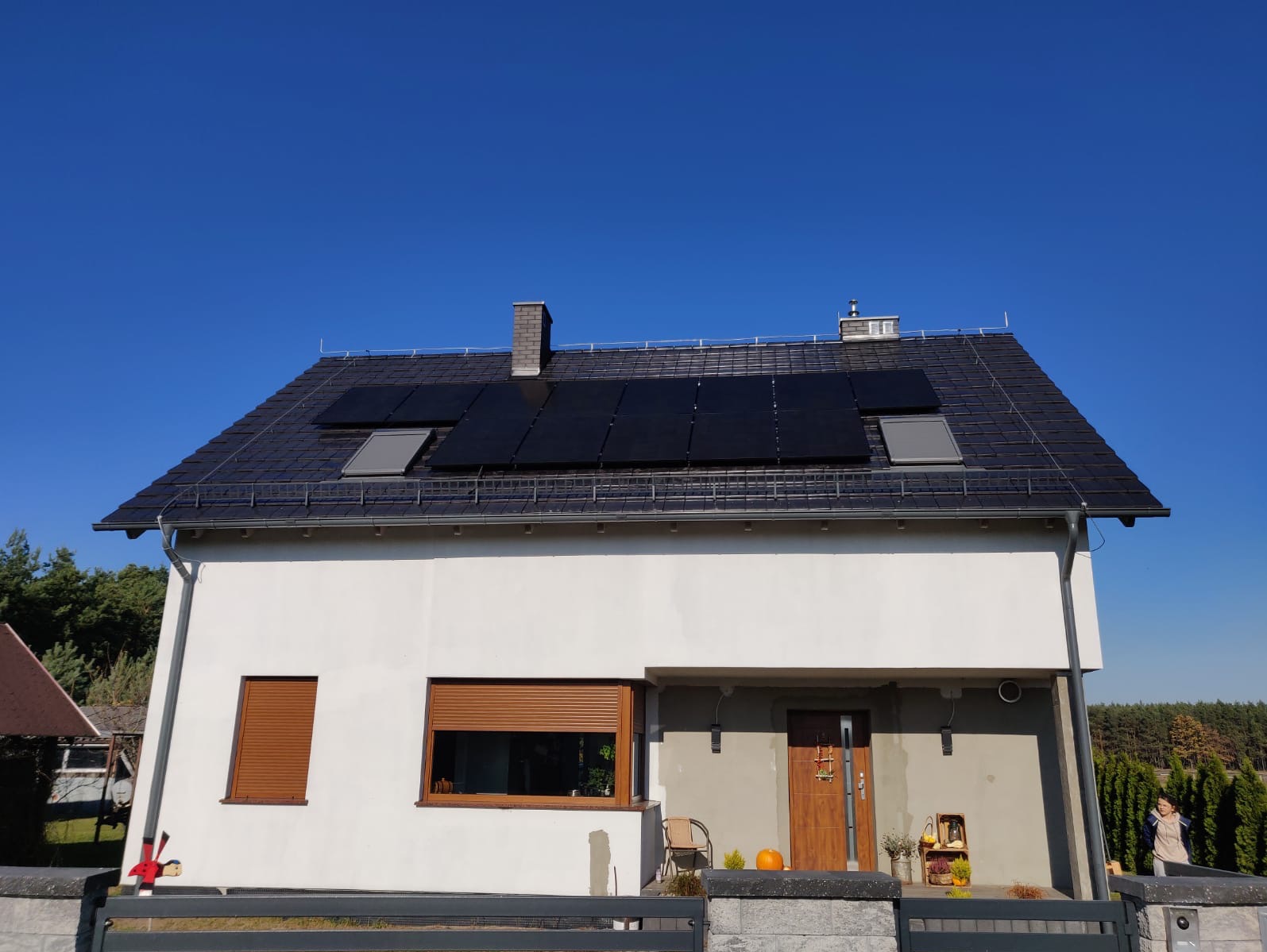 Biały dom jednorodzinny z ciemną dachówką i panelami słonecznymi na dachu, widok z przodu, błękitne niebo w tle.