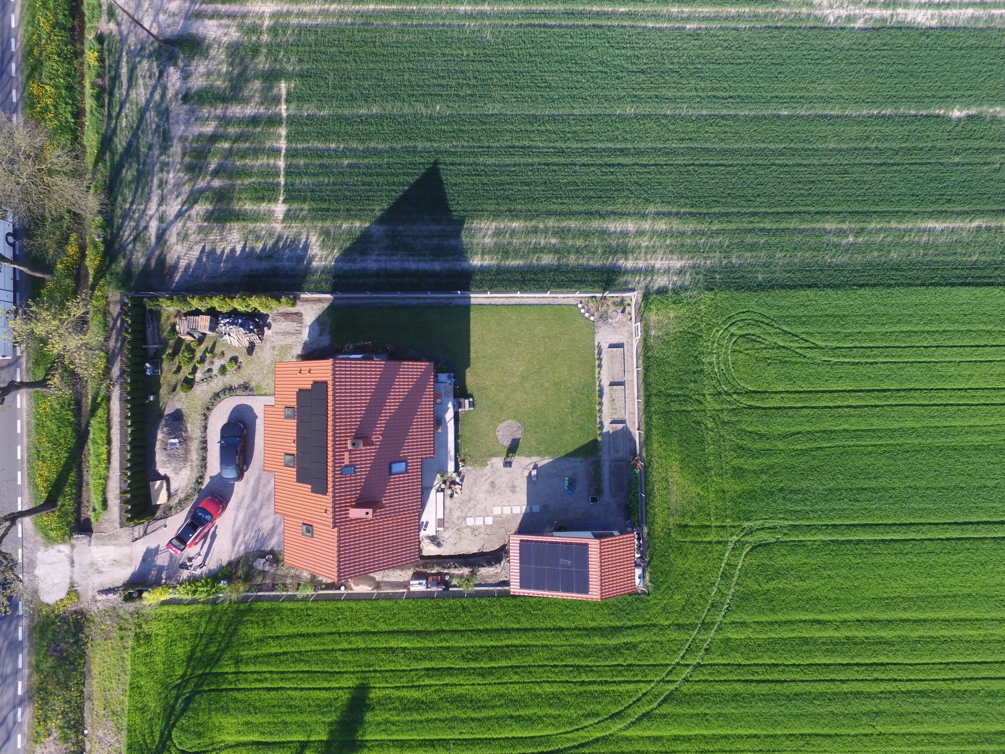 Widok z lotu ptaka na dom z panelami słonecznymi na dachu, obok budynek gospodarczy również z panelami, otoczony zielonym polem i ogrodem, w tle widoczna droga.