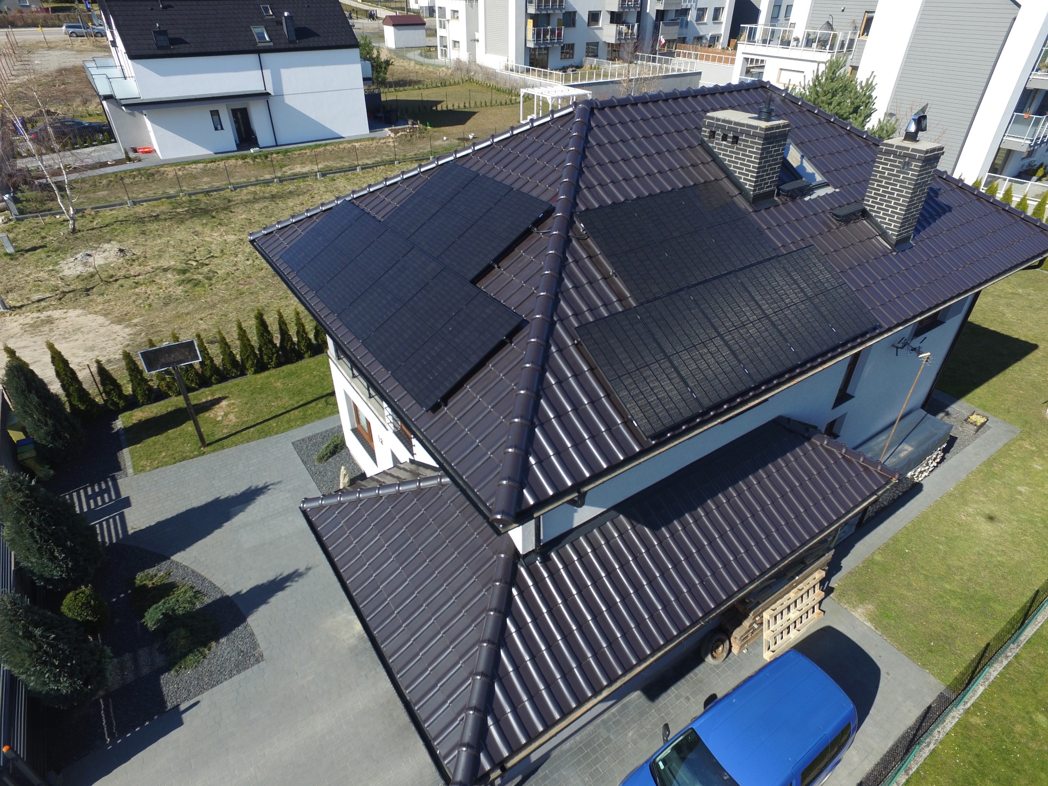 Widok z góry na dom jednorodzinny z brązowym dachem pokrytym panelami słonecznymi; na podjeździe stoi niebieski samochód dostawczy.