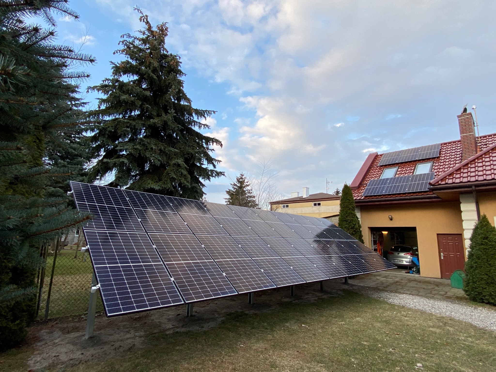 Instalacja fotowoltaiczna na gruncie, panele ustawione pod kątem, dom z panelami na dachu w tle, widoczny samochód w garażu, pochmurne niebo.