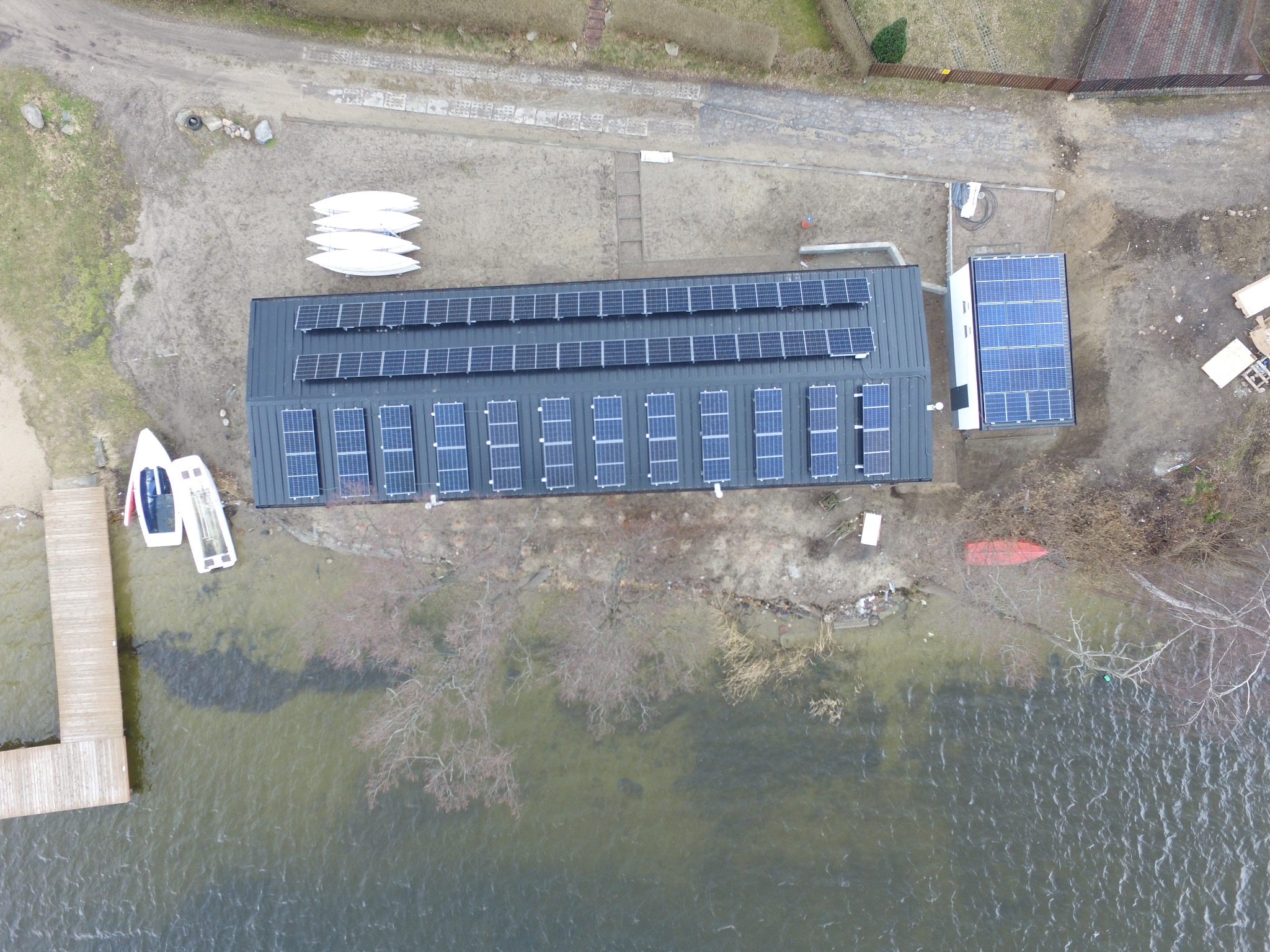 Widok z lotu ptaka na budynek z panelami słonecznymi w Gdyni, położony nad brzegiem jeziora, obok pomostu i kilku kajaków.