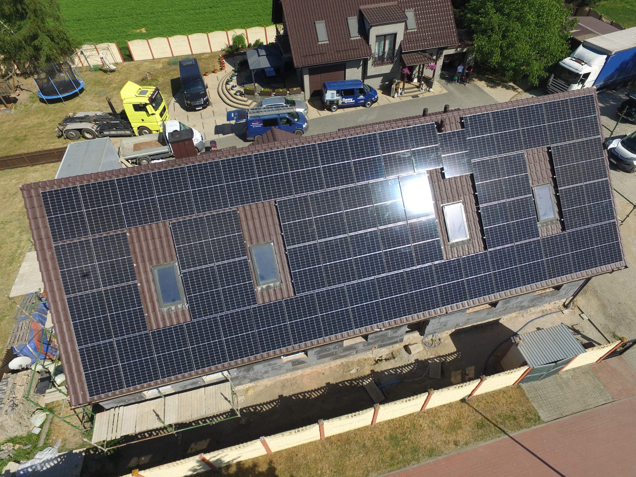 Widok z drona na dom z panelami fotowoltaicznymi na brązowym dachu, z widocznymi oknami dachowymi, częściowo oświetlonymi przez słońce. Na posesji zaparkowane różne pojazdy, w tym samochody...