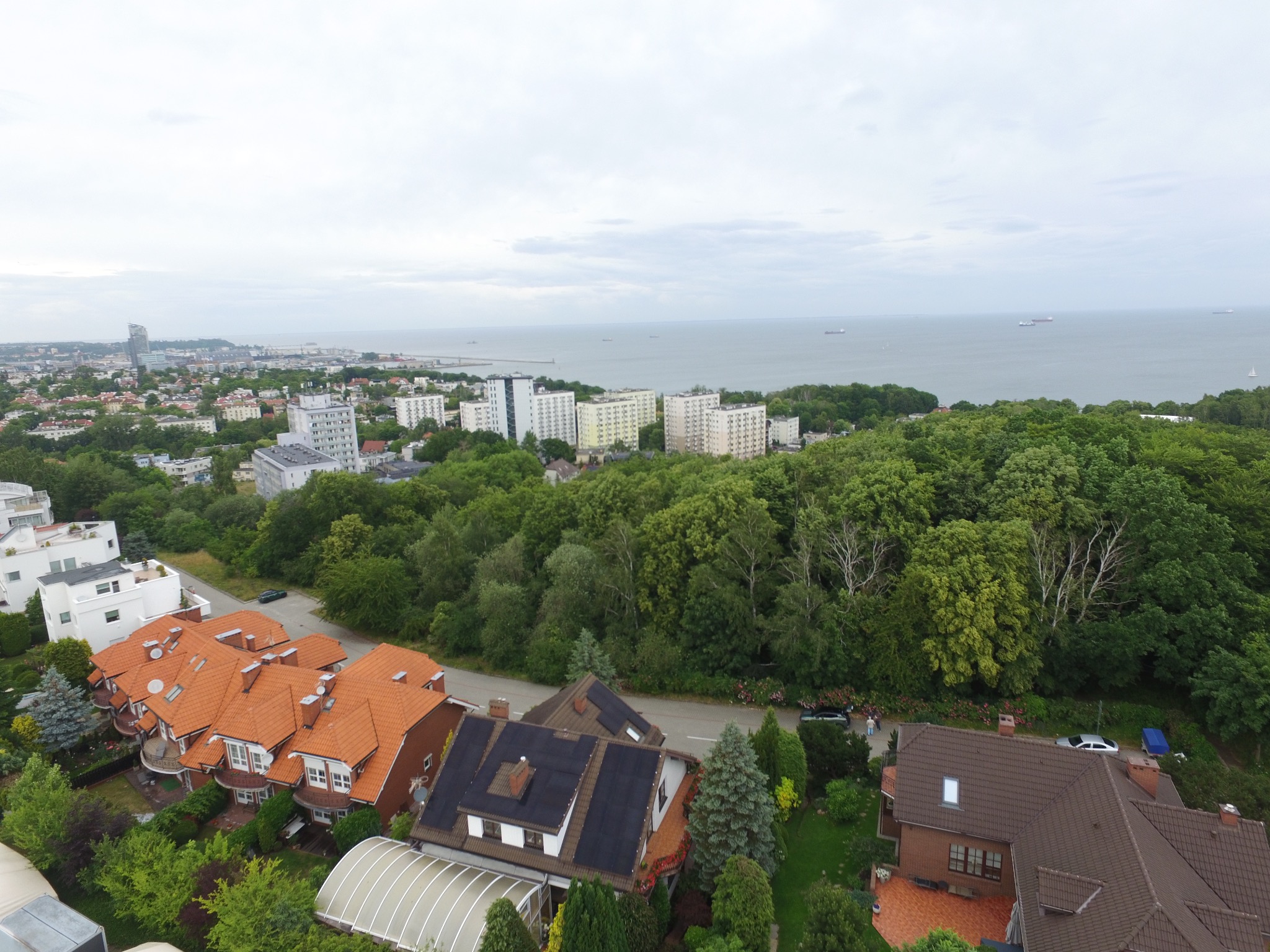Widok z góry na domy z panelami słonecznymi na dachu, w tle zabudowa miejska, morze i pochmurne niebo.