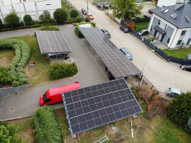 Widok z góry na instalację fotowoltaiczną pełniącą funkcję zadaszenia parkingowego, z czerwonym vanem w pobliżu, na tle zabudowy mieszkalnej.