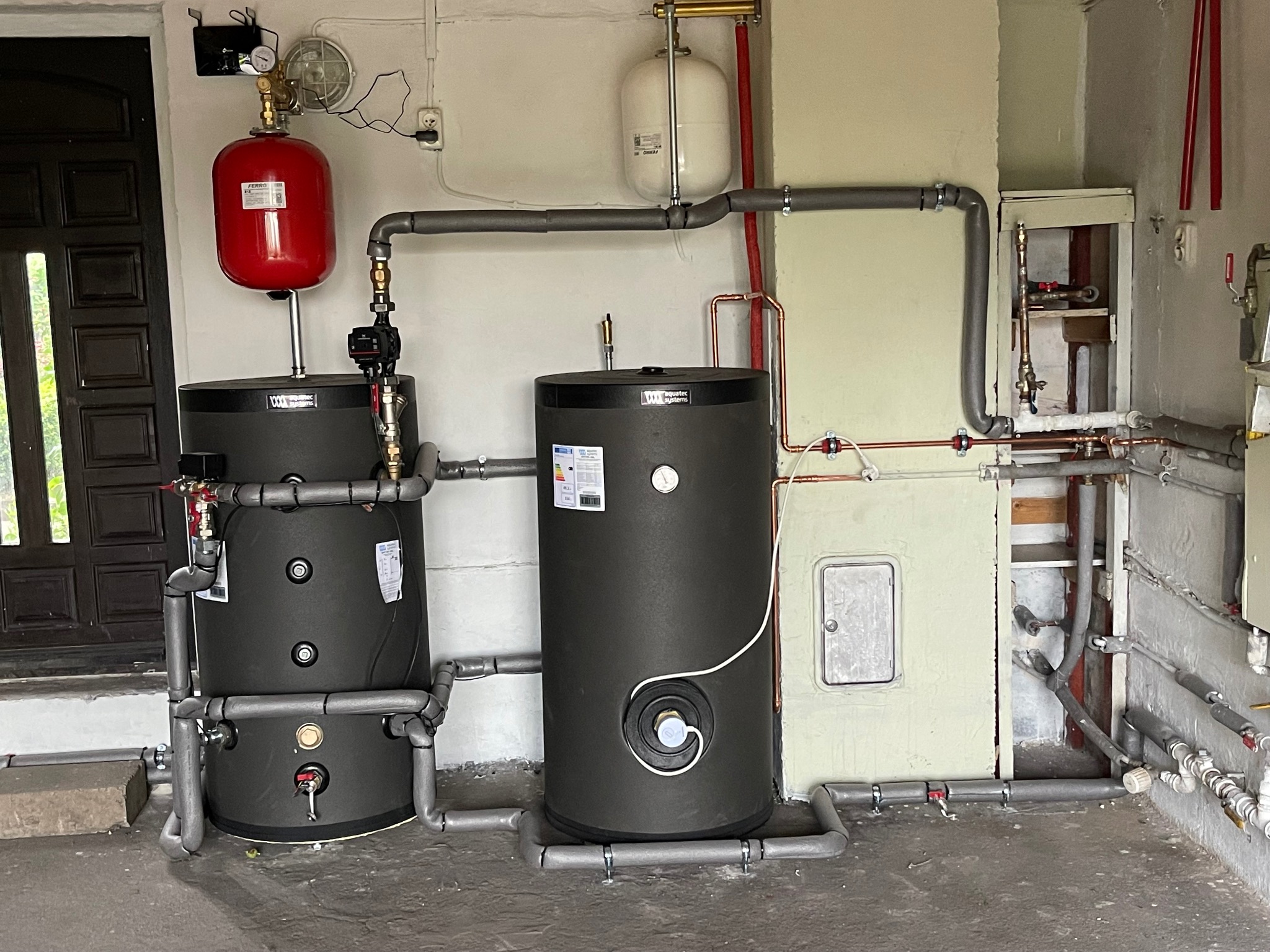 Dwa czarne akumulatory ciepła z czerwoną i białą naczyniem wzbiorczym, połączone rurami w kotłowni z widocznymi elementami instalacji.